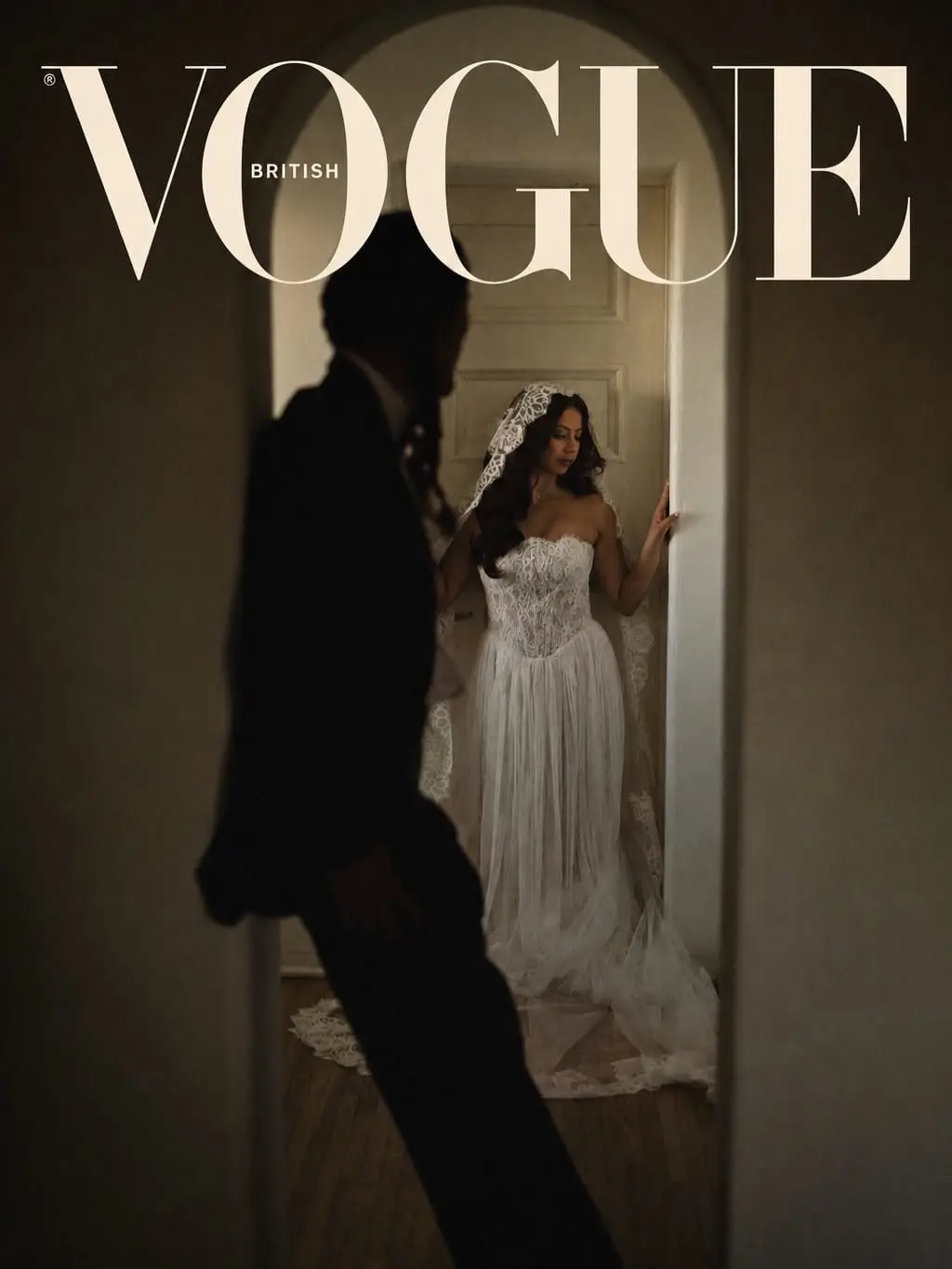 Bride in a strapless lace and tulle gown with a mantilla veil stands framed in a doorway as the groom passes in silhouette, featured in British Vogue.
