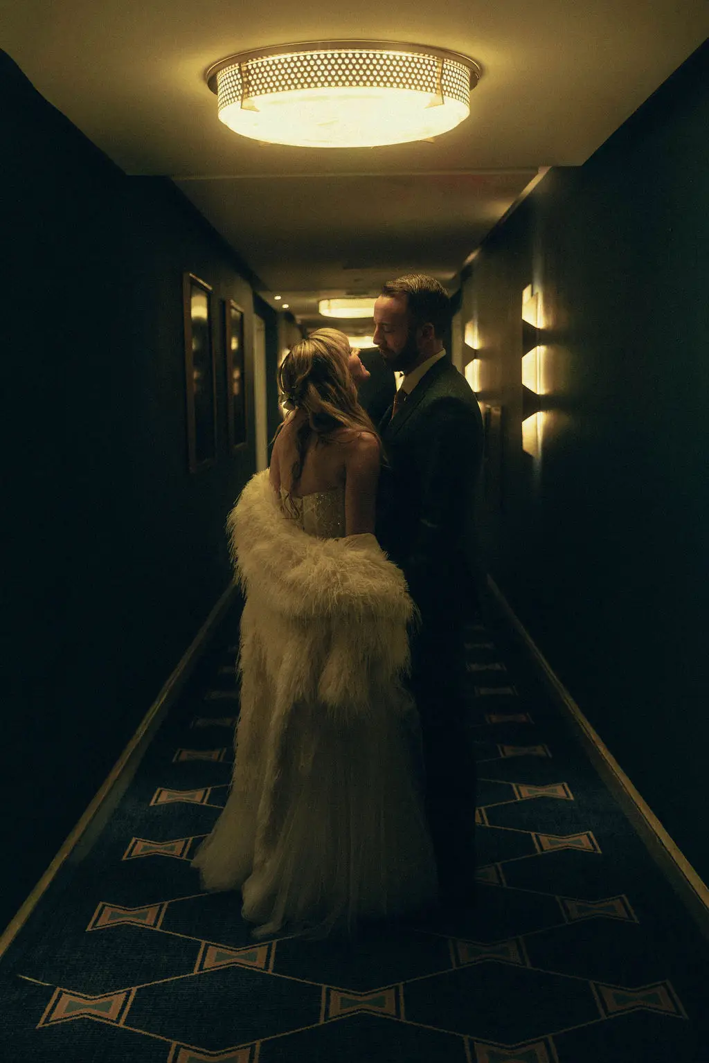Bride and groom embrace in a dim hotel hallway beneath a glowing ceiling light