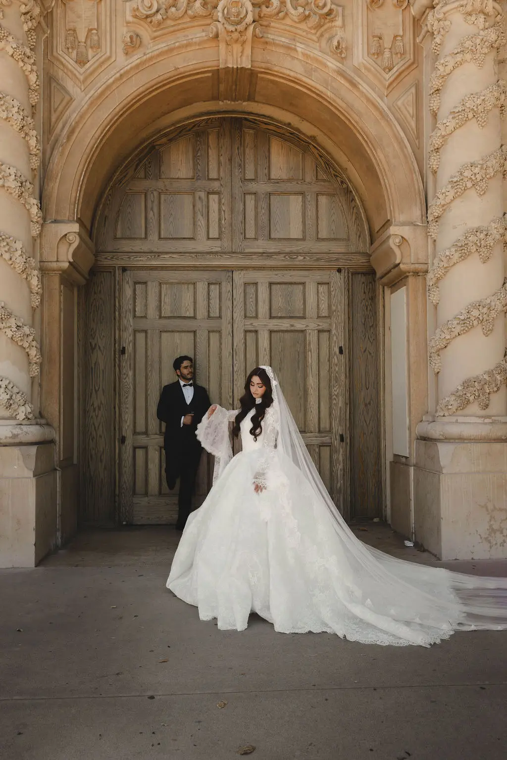 Bride and groom pose under a grand carved stone archway with a long veil trailing