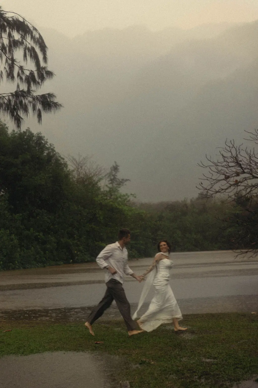 Bride and groom run hand in hand along misty green hills beside water