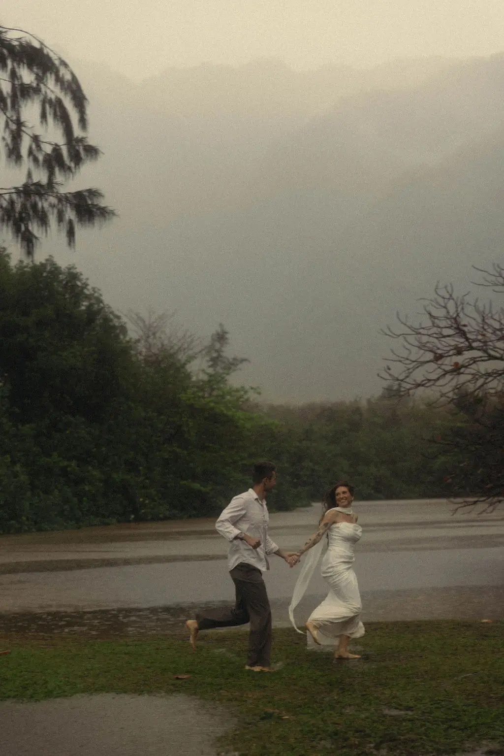 Bride and groom run hand in hand through falling rain before fog-wrapped mountains.
