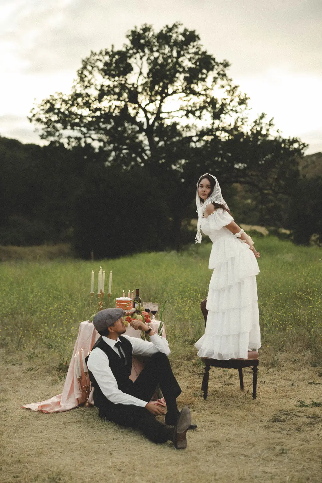 Bride stands over seated groom at a candlelit table set in an open grassy field