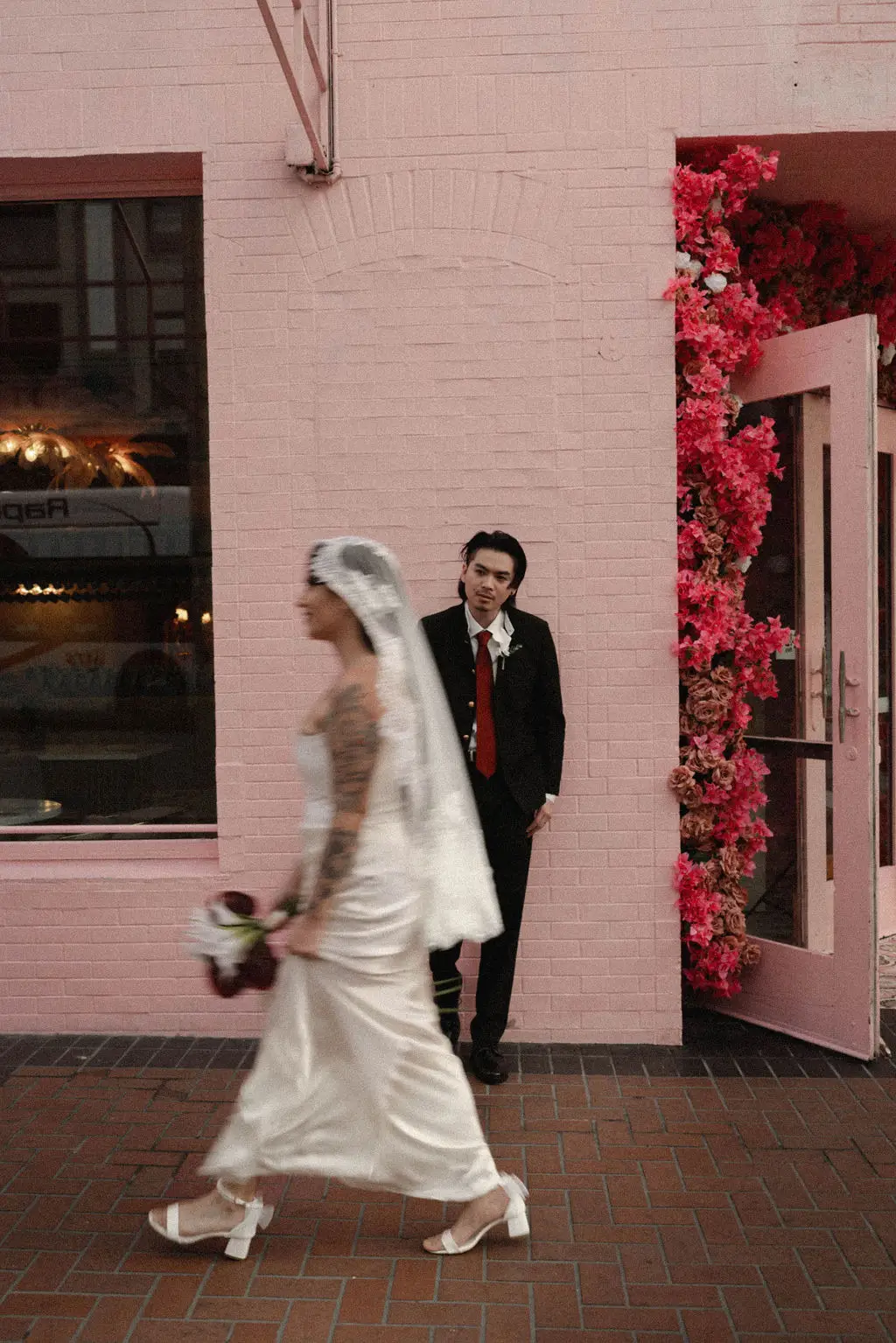 Bride walks past a pink building as groom waits by a bougainvillea-framed doorway