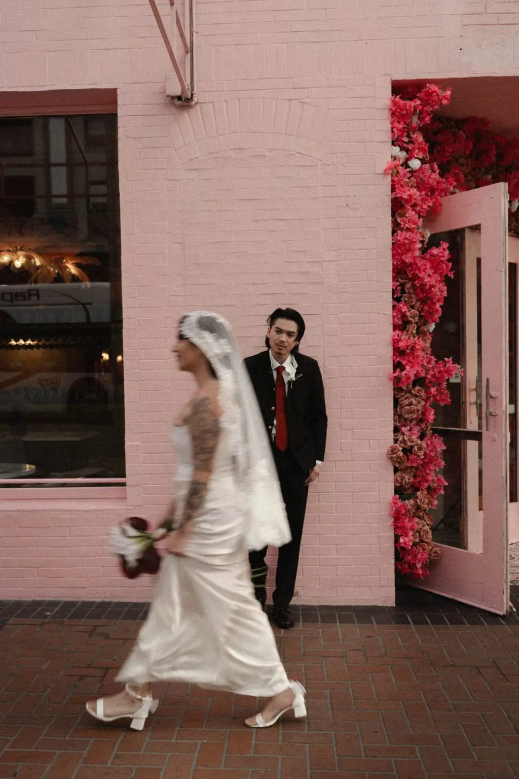 Bride walks past a pink building as groom waits by a bougainvillea-framed doorway
