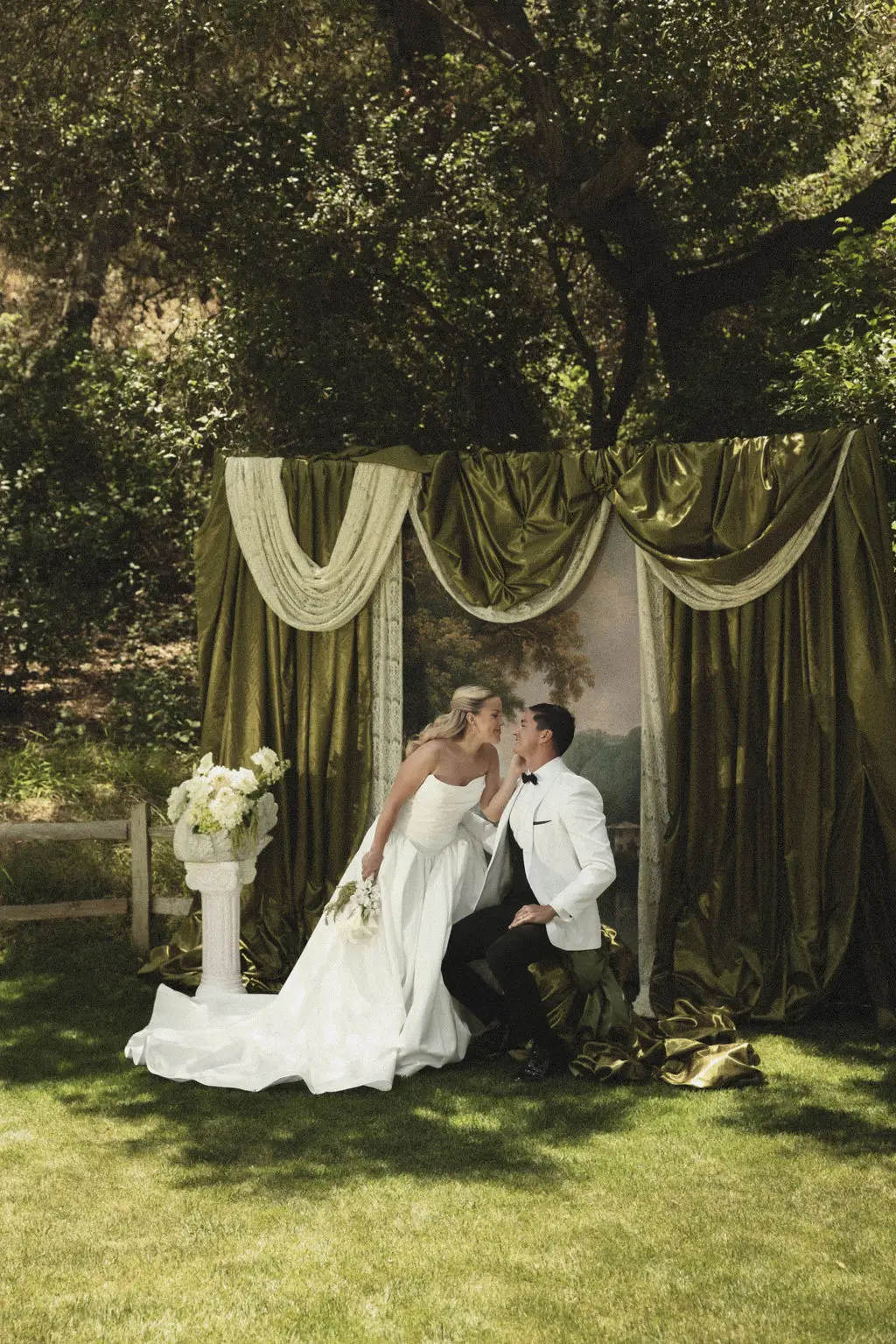 Bride and groom kiss before a draped green velvet altar backdrop on a garden lawn