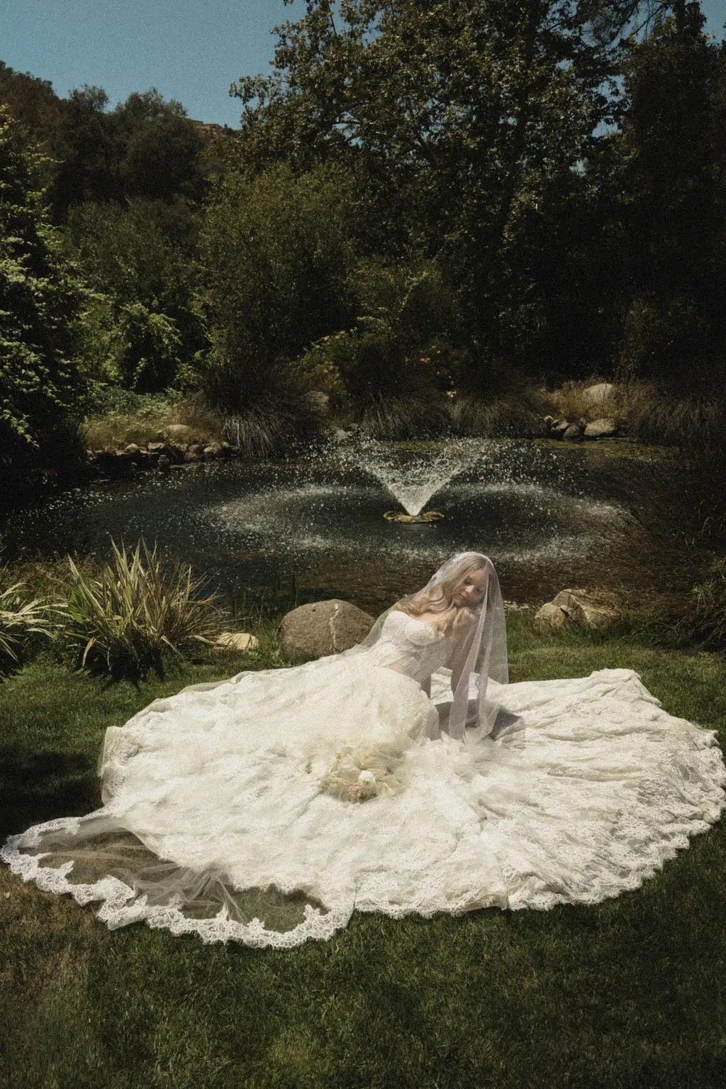 Bride reclines with her gown spread across grass before a garden pond and fountain