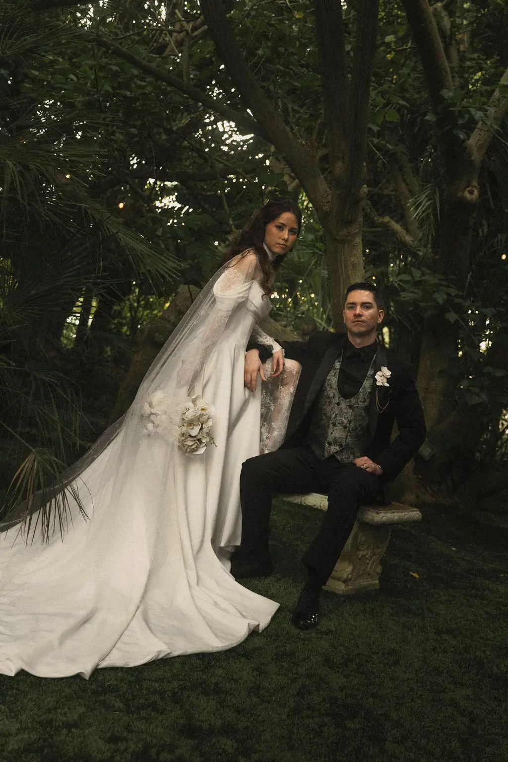 Bride stands beside seated groom on a stone bench in a lush shadowed garden
