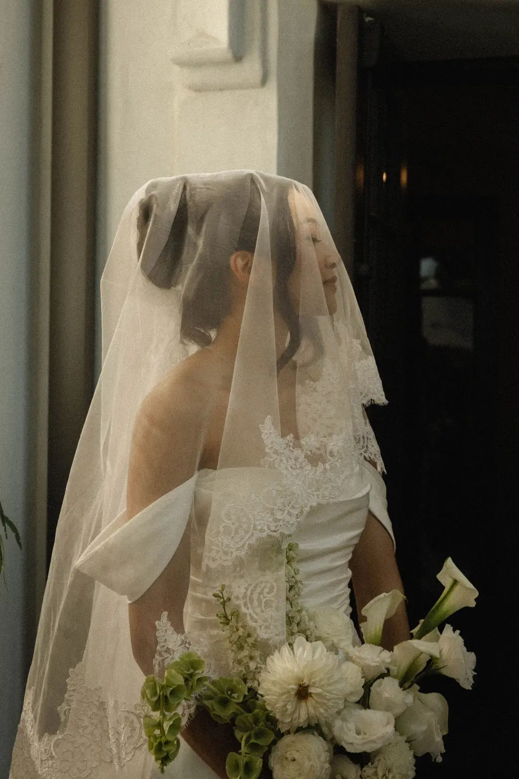 Bride in a lace-edged veil holds a white bouquet, face softly obscured by sheer tulle