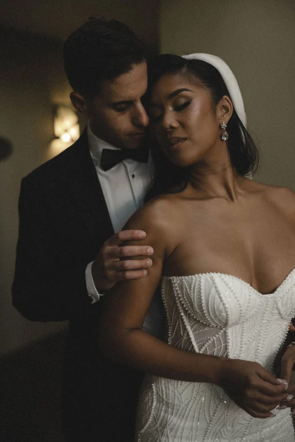 Groom in tuxedo embraces bride from behind in warm interior light, her eyes closed serenely