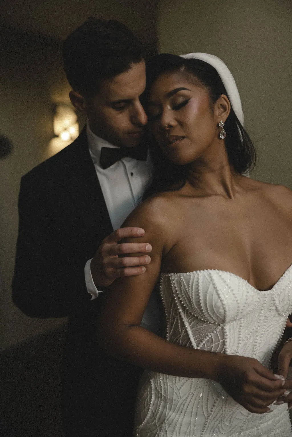 Groom in tuxedo embraces bride from behind in warm interior light, her eyes closed serenely