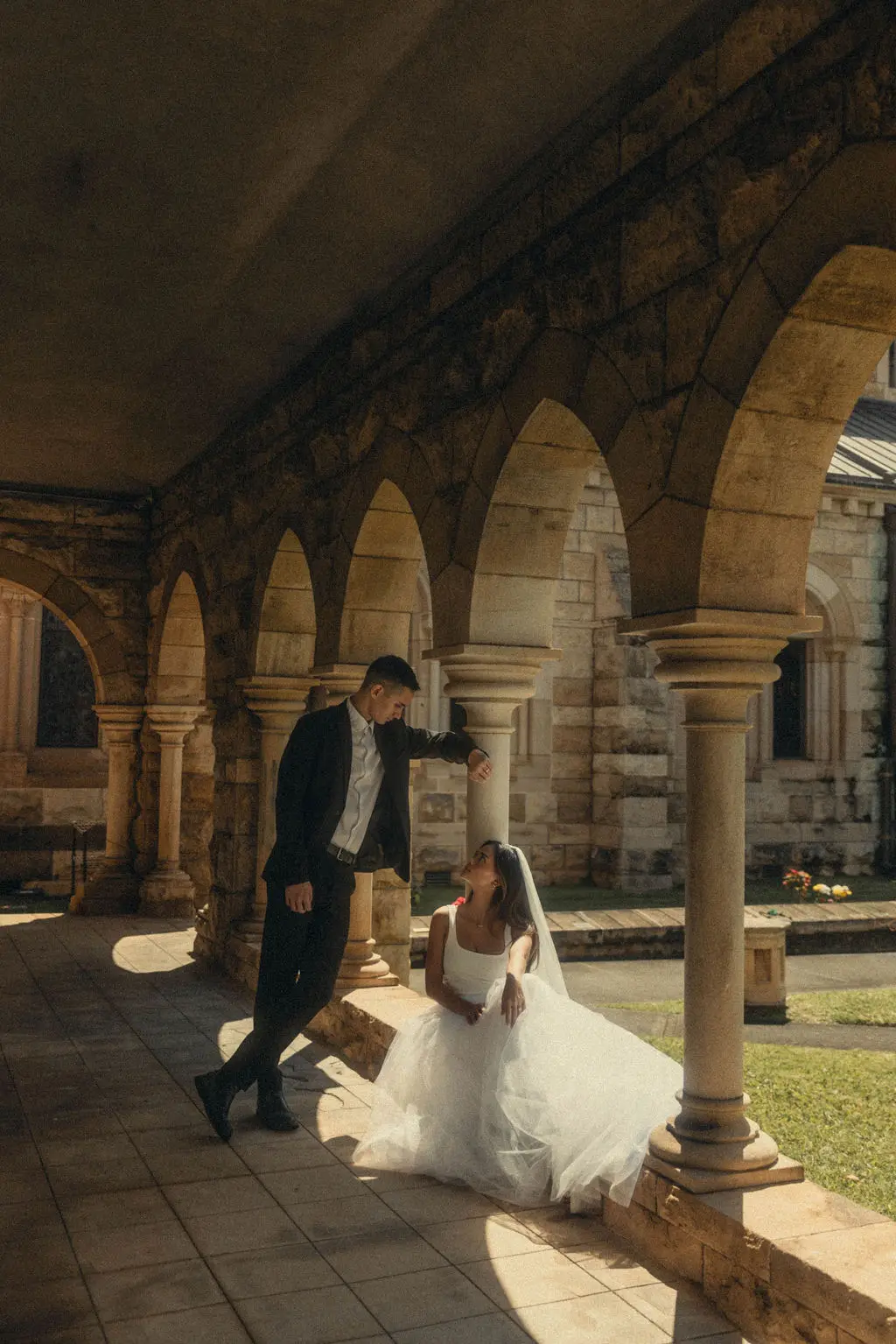 Groom leans toward seated bride in the arched sandstone cloister of a cathedral courtyard