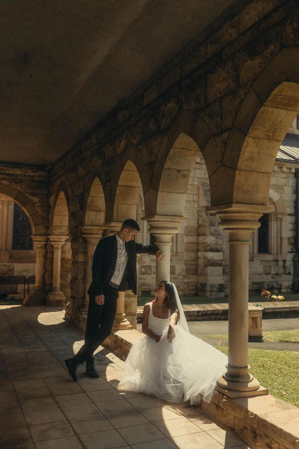 Groom leans toward seated bride in the arched sandstone cloister of a cathedral courtyard