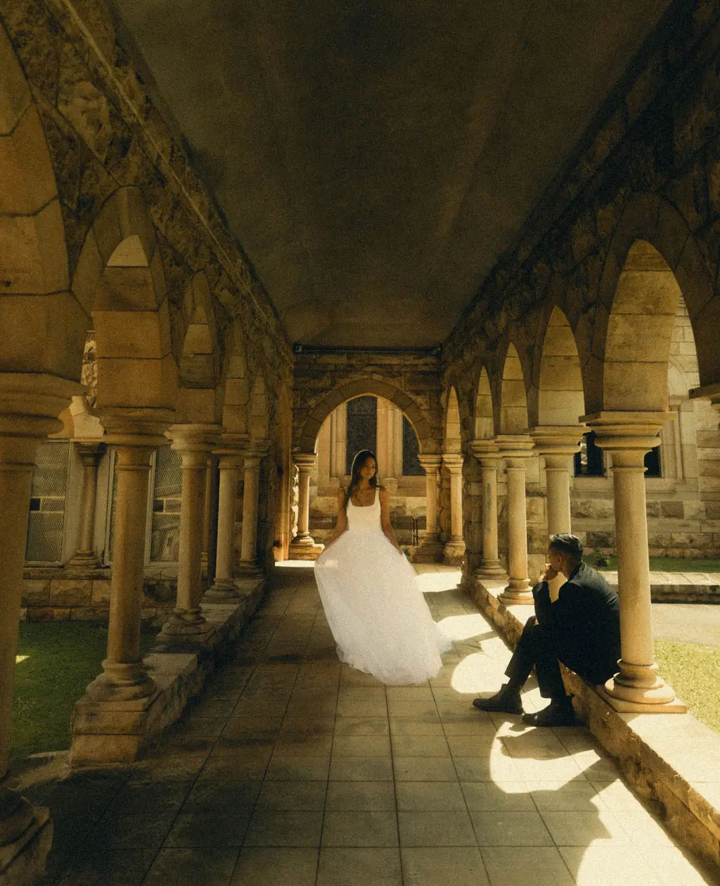 Bride in a flowing gown within a sunlit arched sandstone cloister as the groom rests nearby