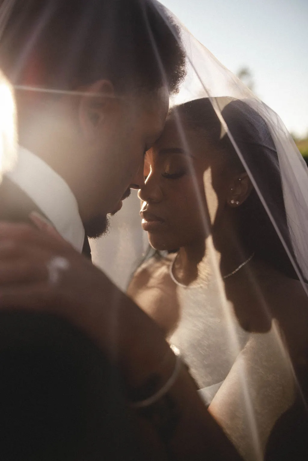 Bride and groom press foreheads together beneath a backlit veil in golden sunlight