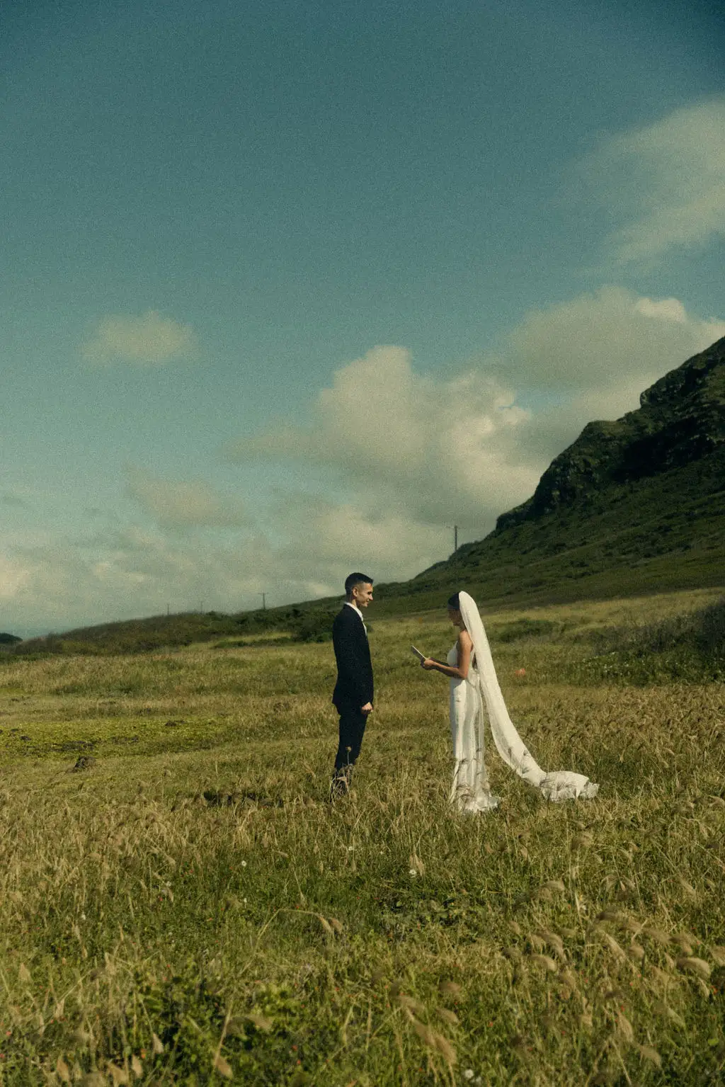 Groom reads vows to bride in a windswept green field beneath a mountain ridge and open sky