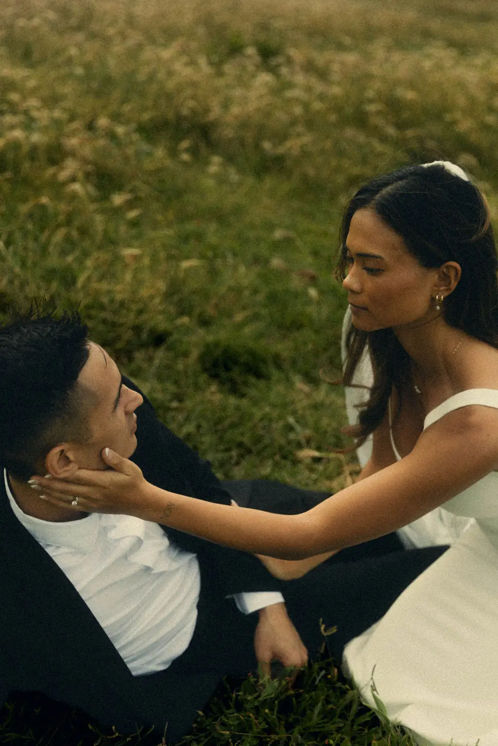 Bride cradles the groom's face as they sit together in a sunlit meadow of tall grass
