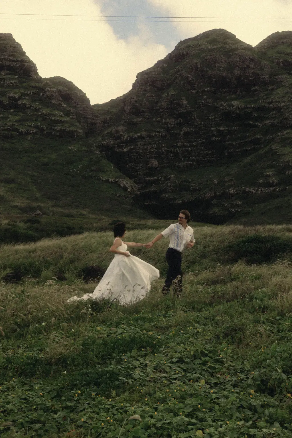 Groom leads bride by the hand through a lush green valley below steep mist-touched mountains