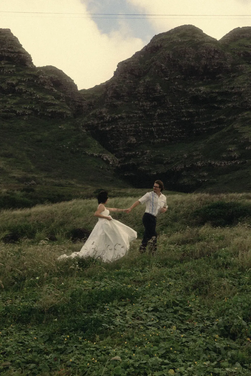 Groom leads bride by the hand through a lush green valley below steep mist-touched mountains