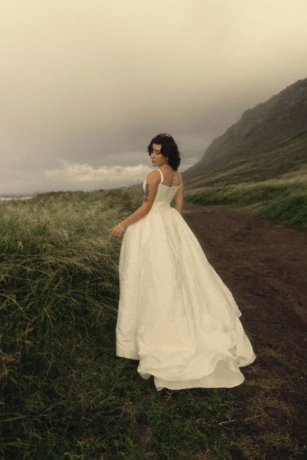 Bride in a full gown glances over her shoulder on a misty coastal path below a green headland