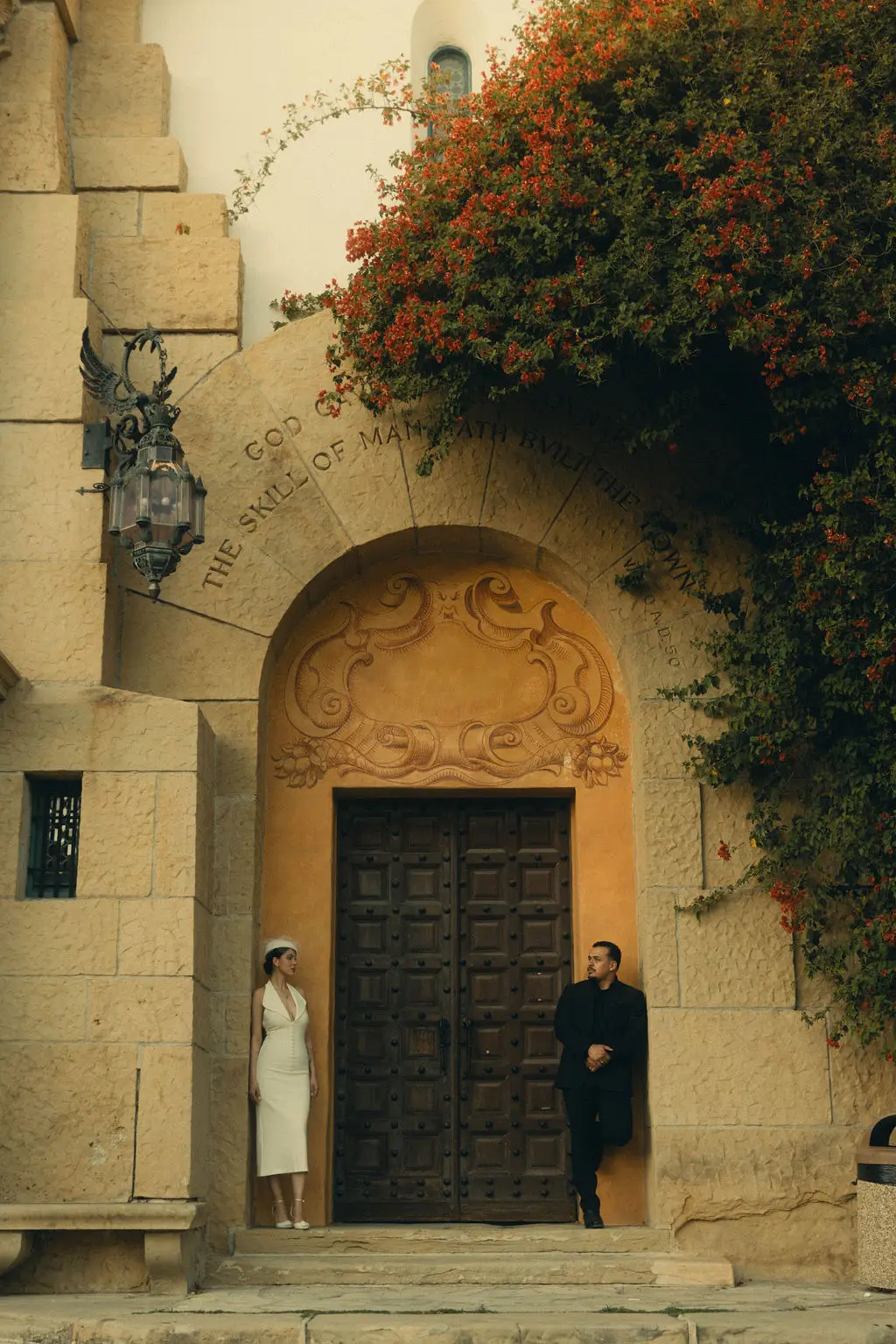 Bride and groom stand apart at a grand carved wooden doorway framed by stone and red blossoms