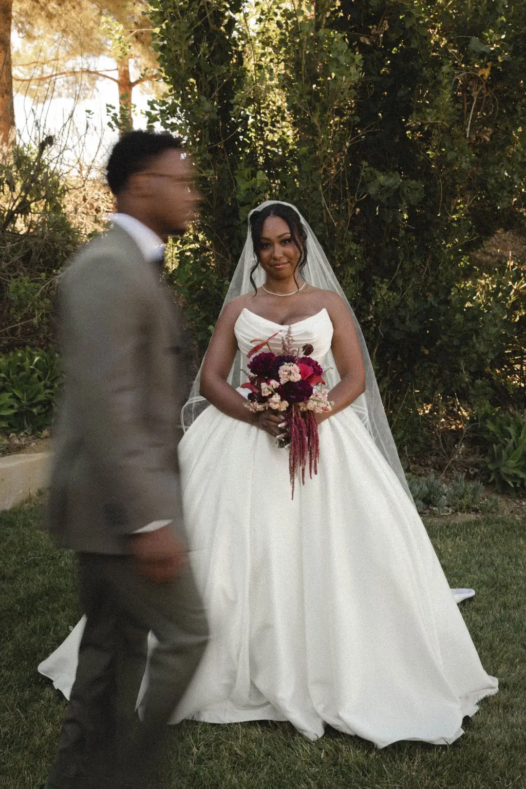 Veiled bride with burgundy bouquet smiles at her groom during a garden first look