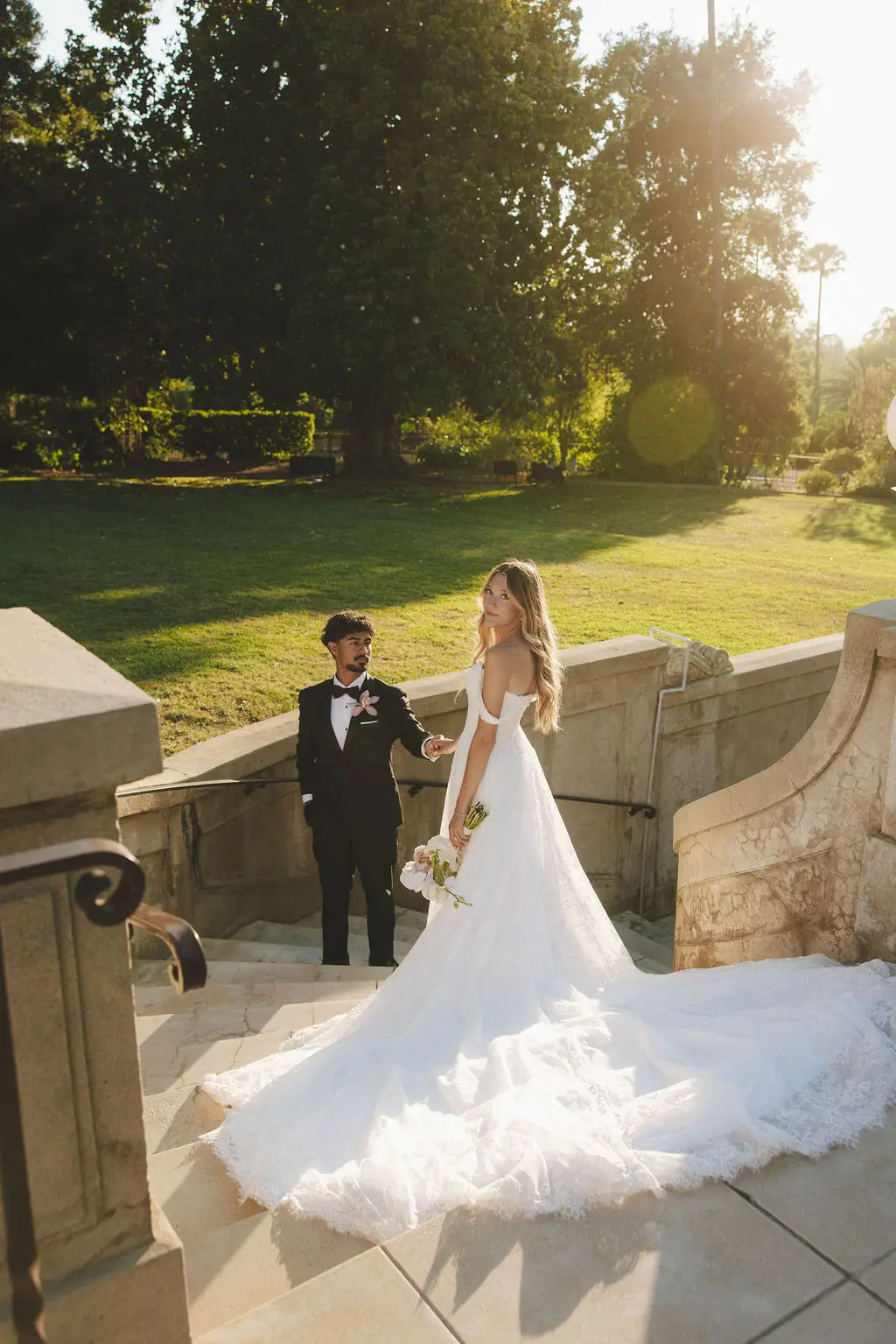 Bride with long train and groom in tuxedo pause on grand stone steps in golden light