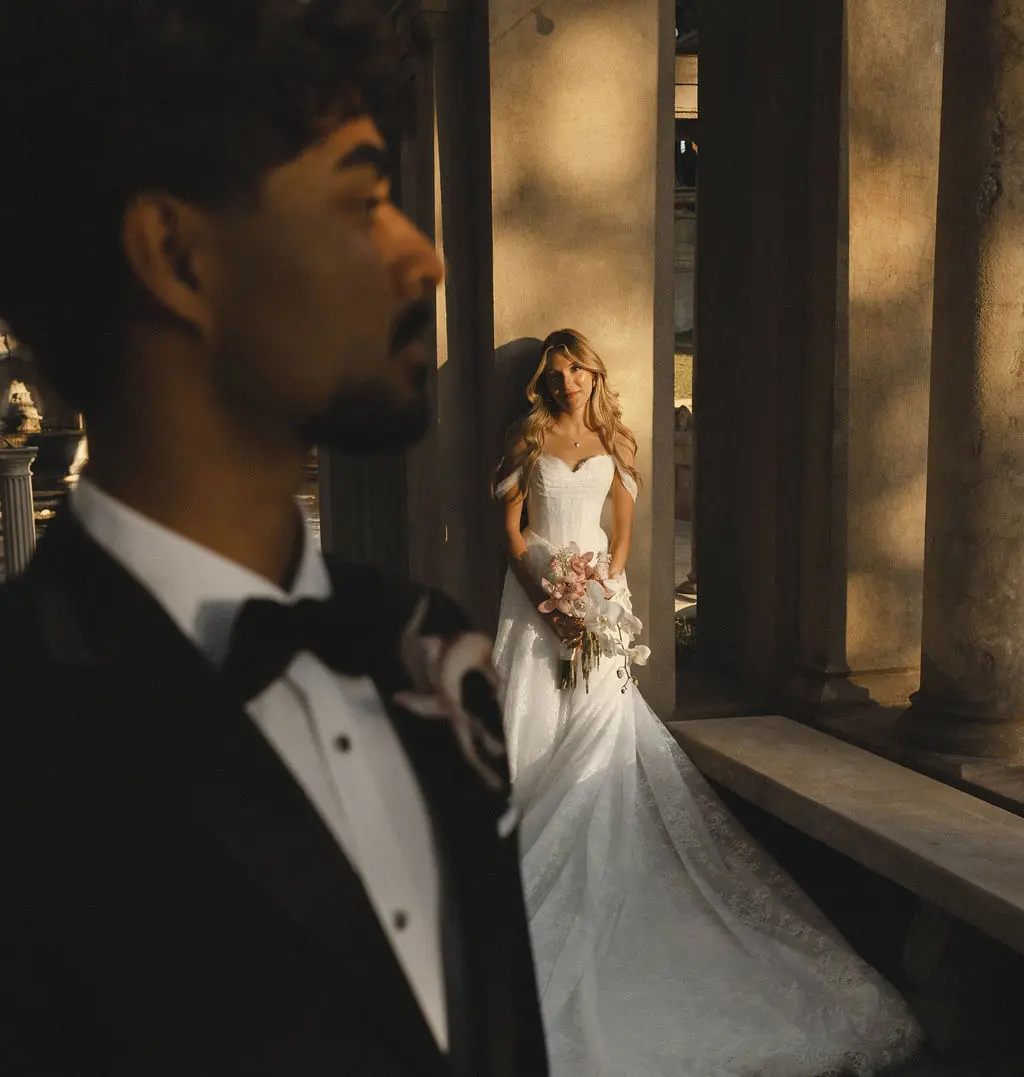 Bride stands among stone columns while the groom looks on in the foreground of a grand interior.