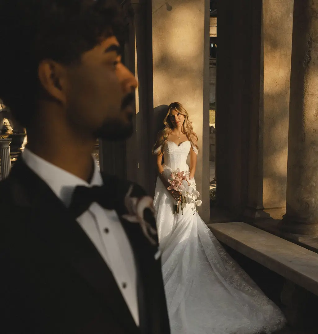 Bride stands among stone columns while the groom looks on in the foreground of a grand interior