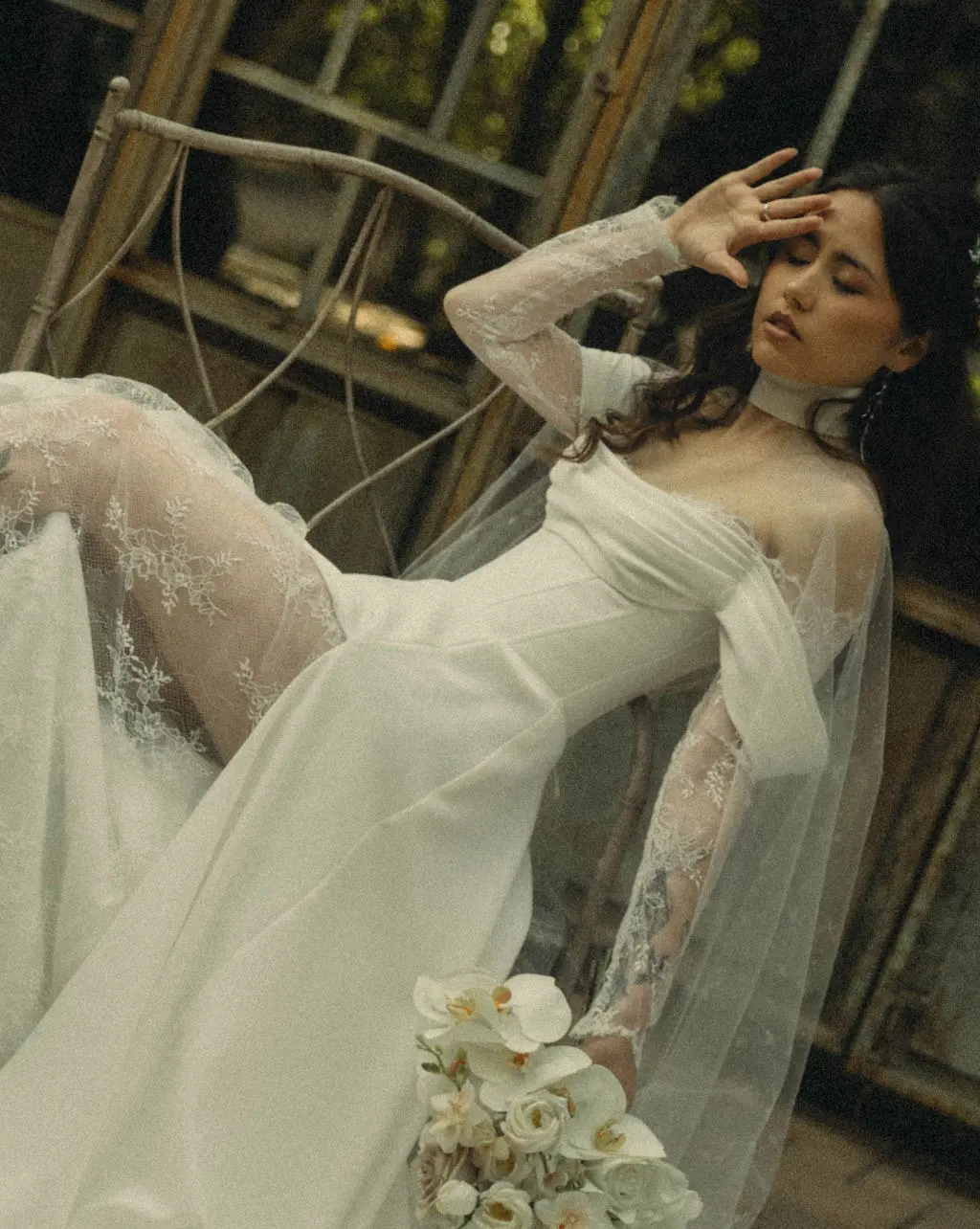 Bride in a sheer lace gown reclines on an iron chair holding a white orchid bouquet