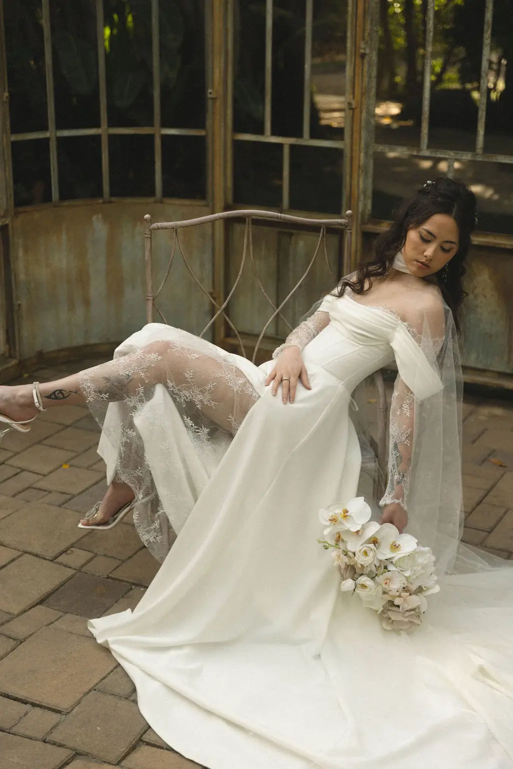Bride in a sheer lace gown reclines on an iron bench with a white orchid bouquet on brick
