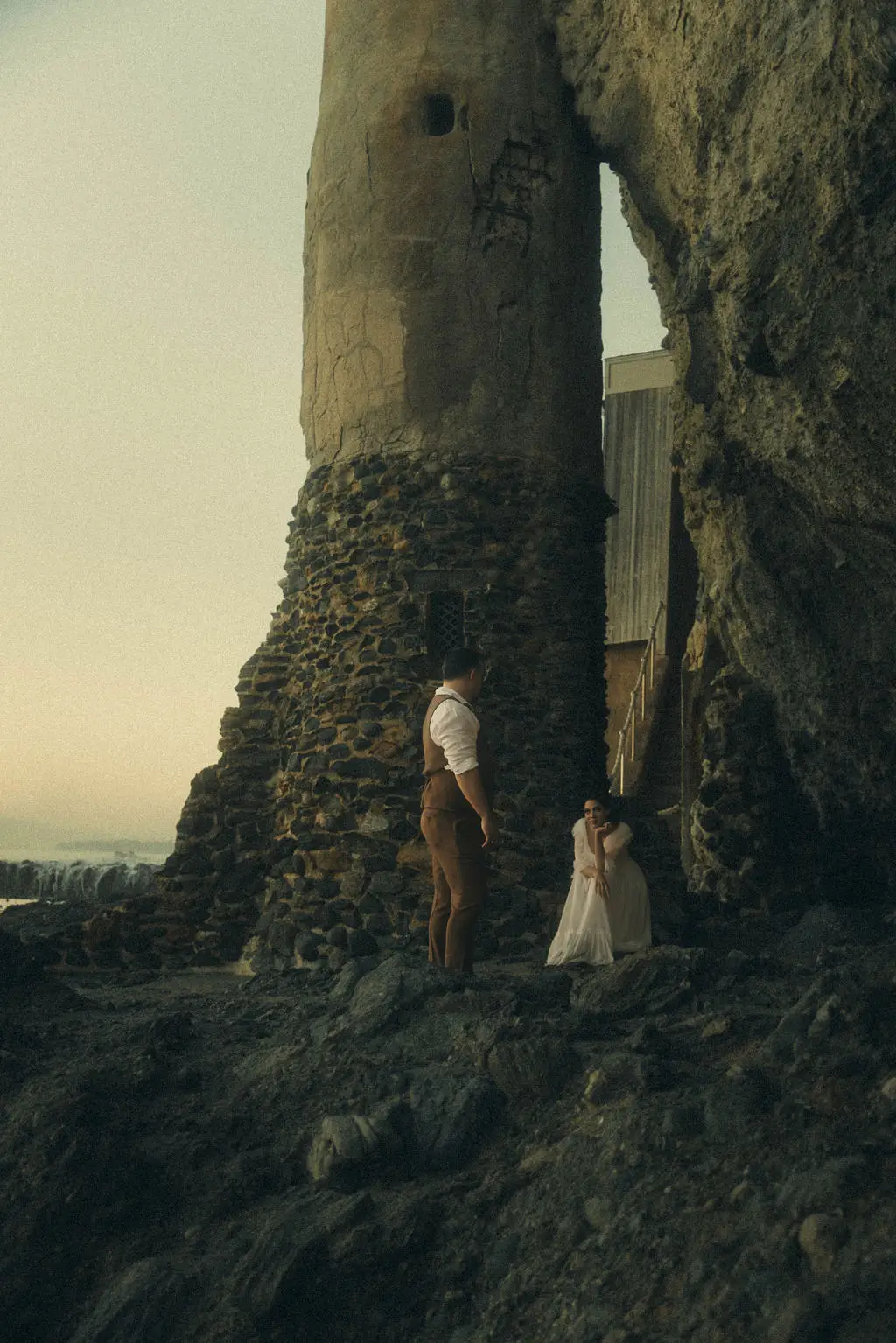 Couple stands small against a towering stone sea stack on a rocky coastline at dusk