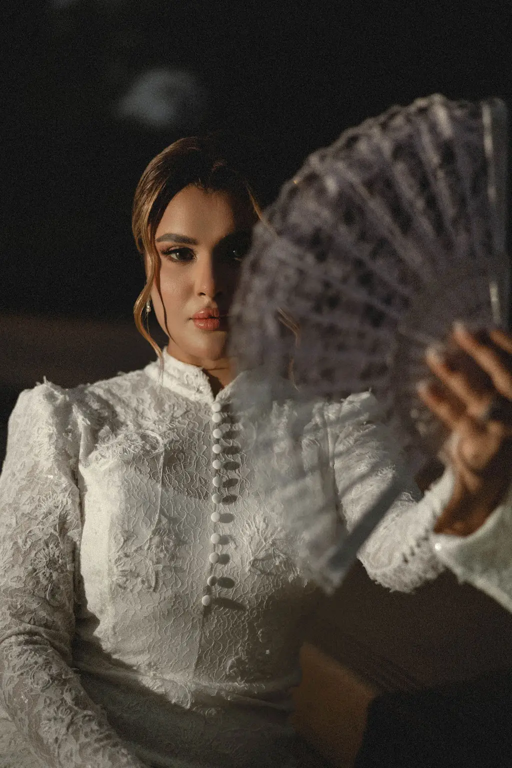 Bride in a high-neck lace gown gazes past a lace hand fan in dramatic low light