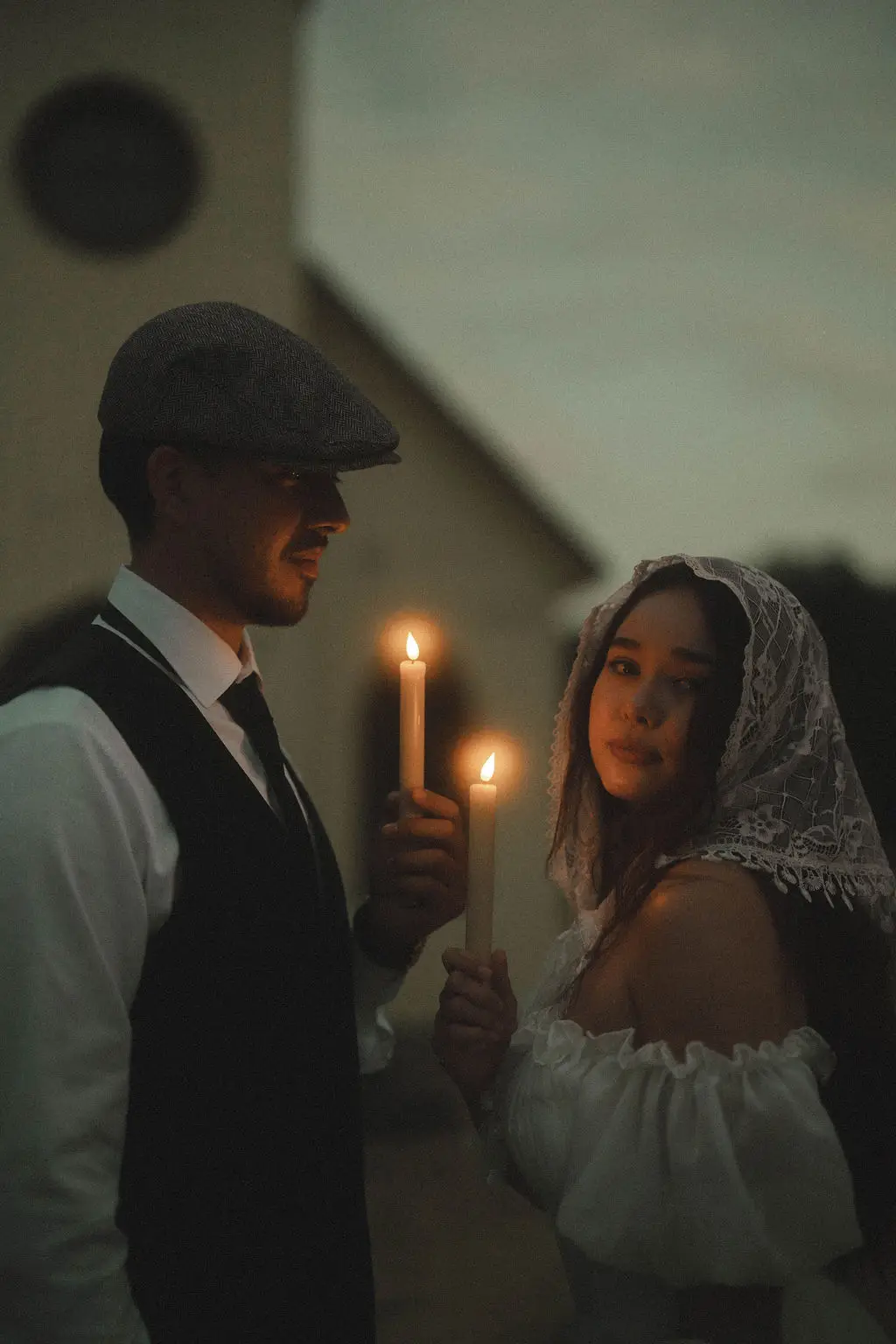 Bride and groom hold lit candles at dusk beside a white chapel wall in vintage styling