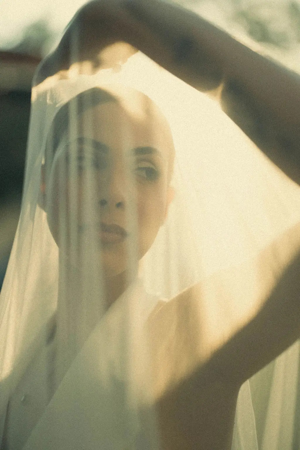 Sunlit bride lifts her sheer veil overhead, her face glowing softly behind the fabric