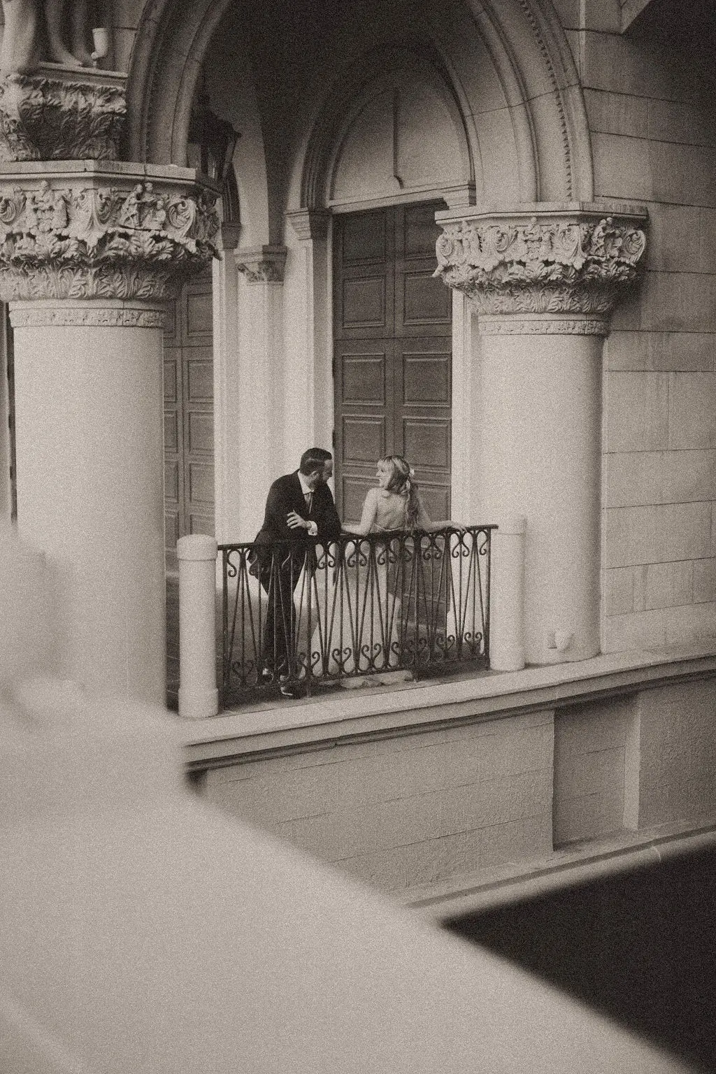 Couple embraces on a grand arched stone balcony seen from across the building in sepia