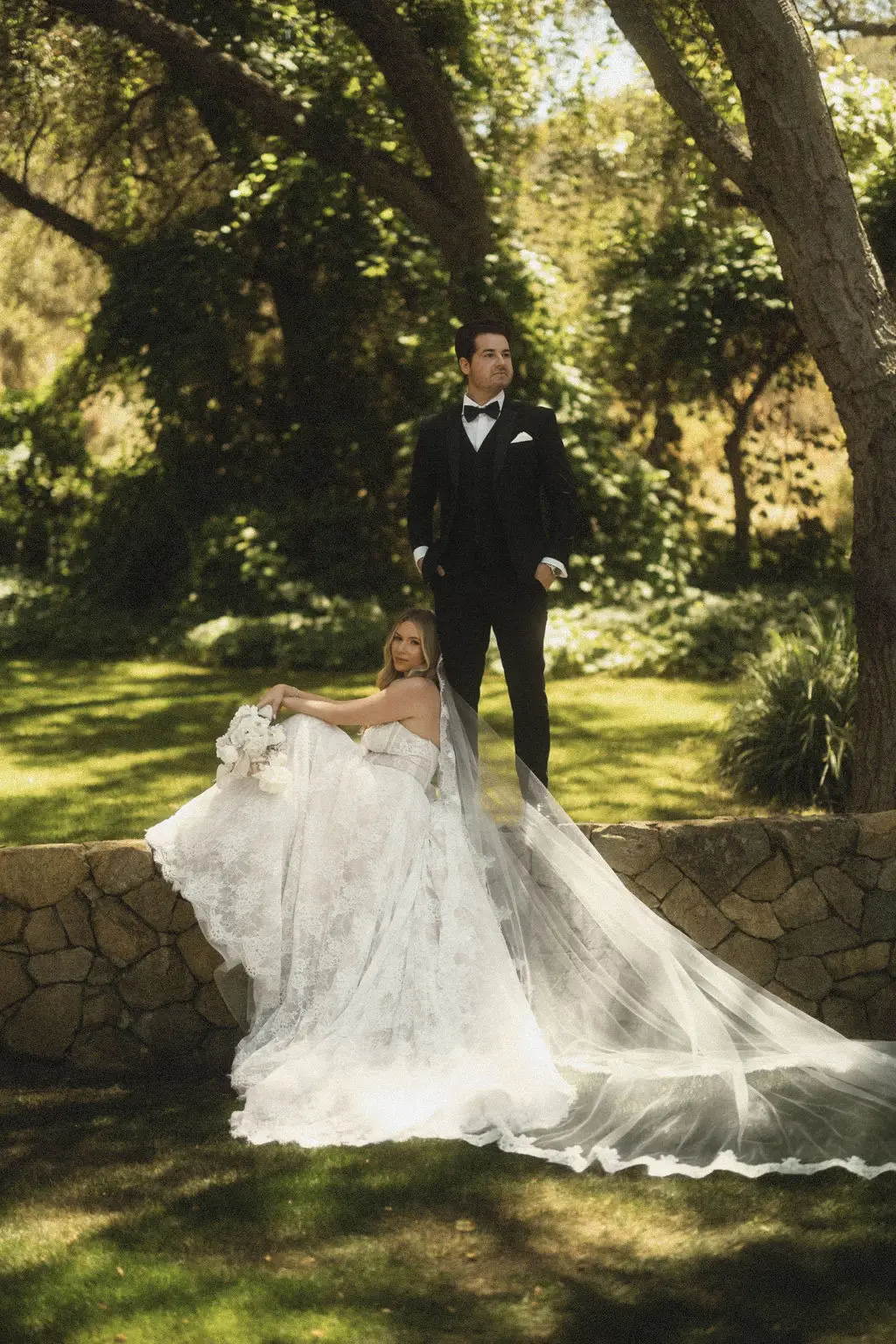 Groom stands on a stone wall as the bride sits below with her veil trailing across sunlit grass