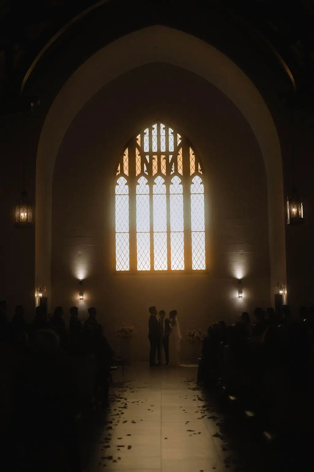 Bride and groom exchange vows before a glowing gothic arched window inside a dim chapel