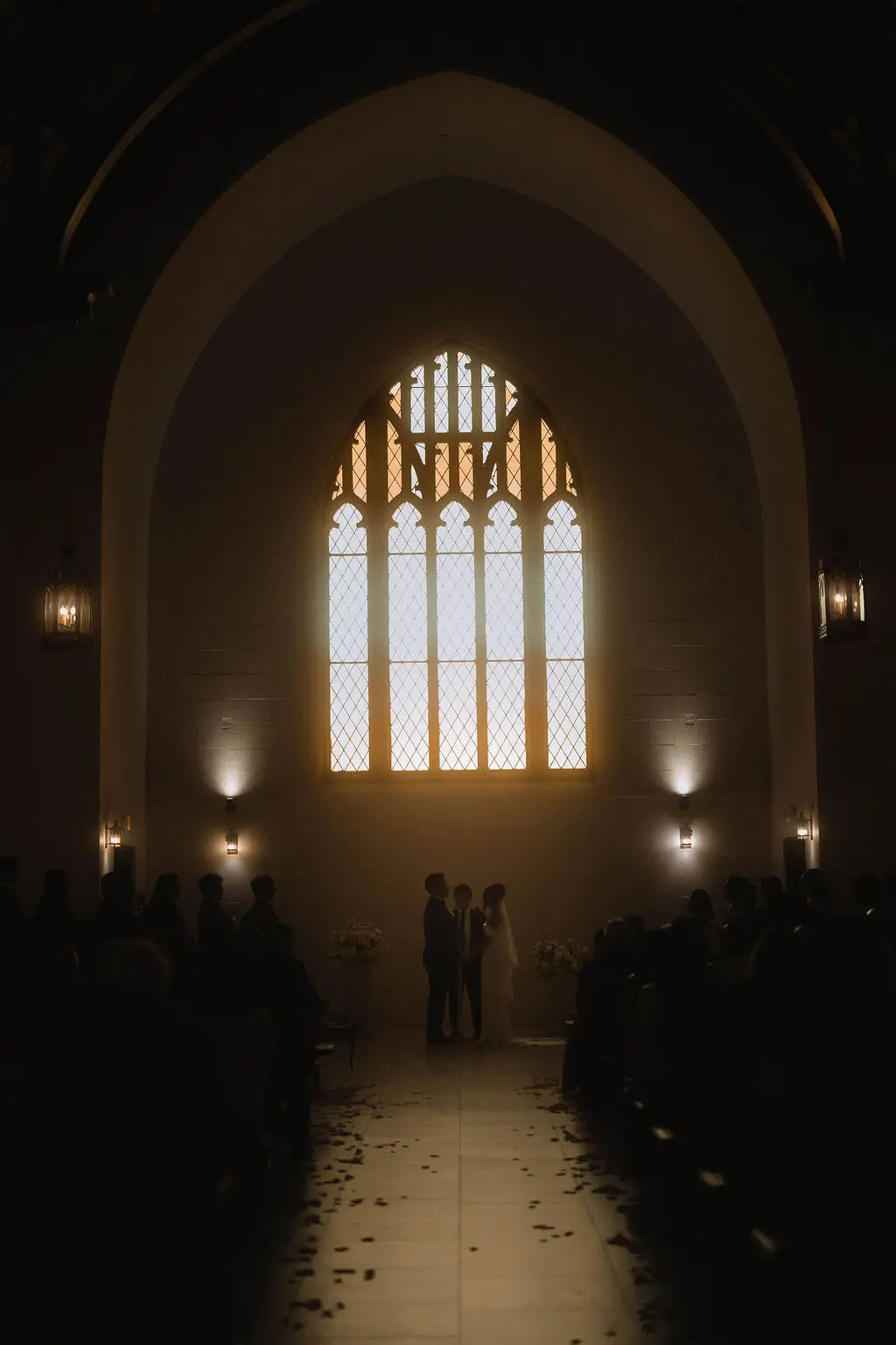 Bride and groom exchange vows before a glowing gothic arched window inside a dim chapel