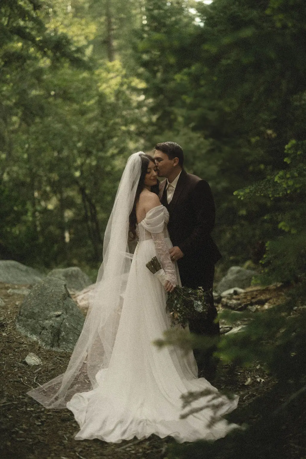 Groom kisses veiled bride's forehead among mossy boulders and forest greenery at a woodland wedding