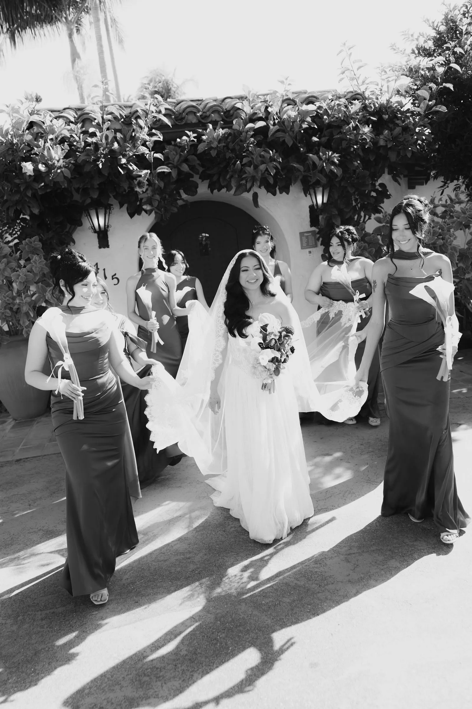 Black-and-white photograph of the bride walking with her bridesmaids through a sunlit courtyard at Casa Romantica, her lace veil catching the coastal breeze.