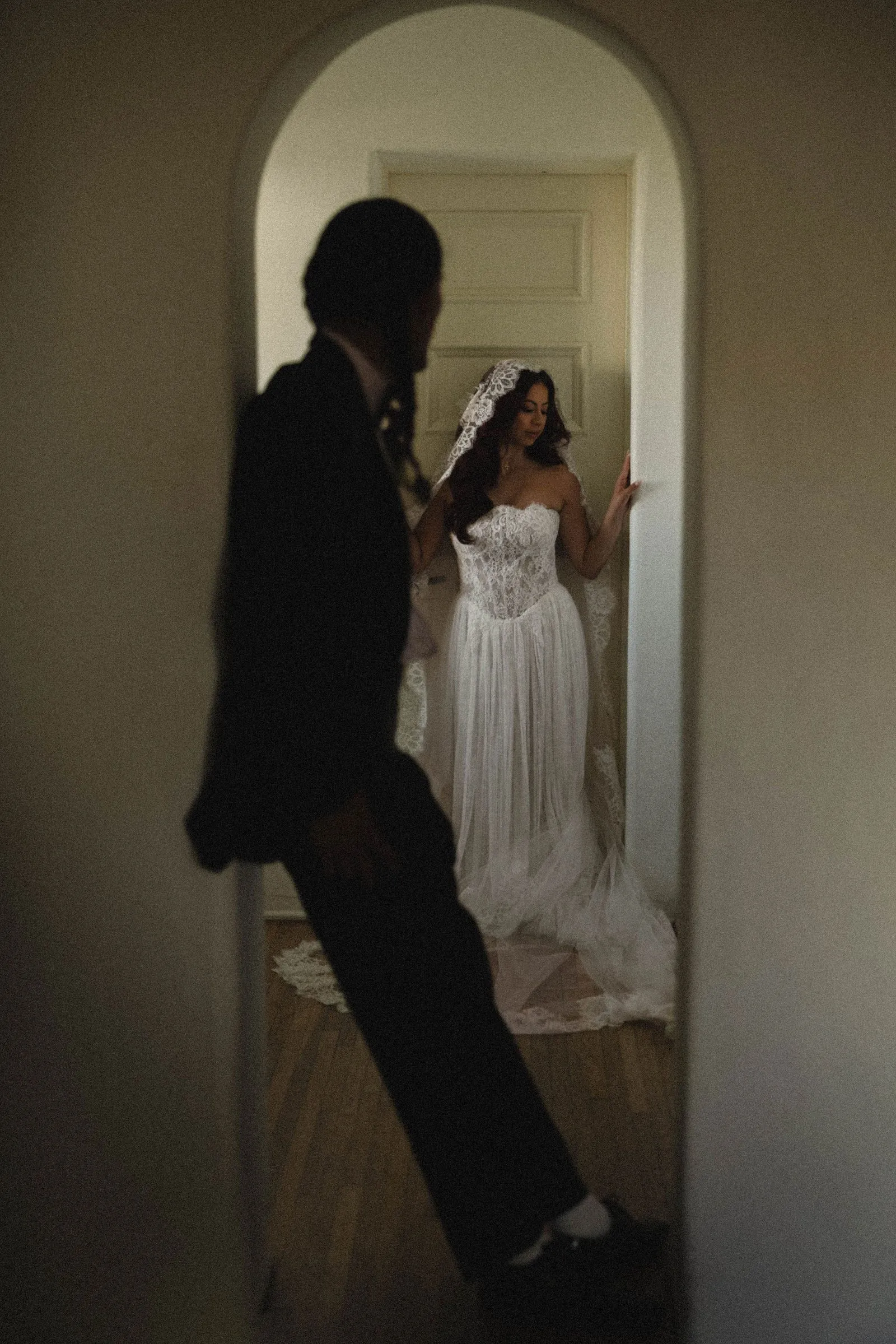 Bride in a white arched doorway at Casa Romantica while the groom stands in silhouette, a quiet moment of Spanish Colonial architecture and soft light.