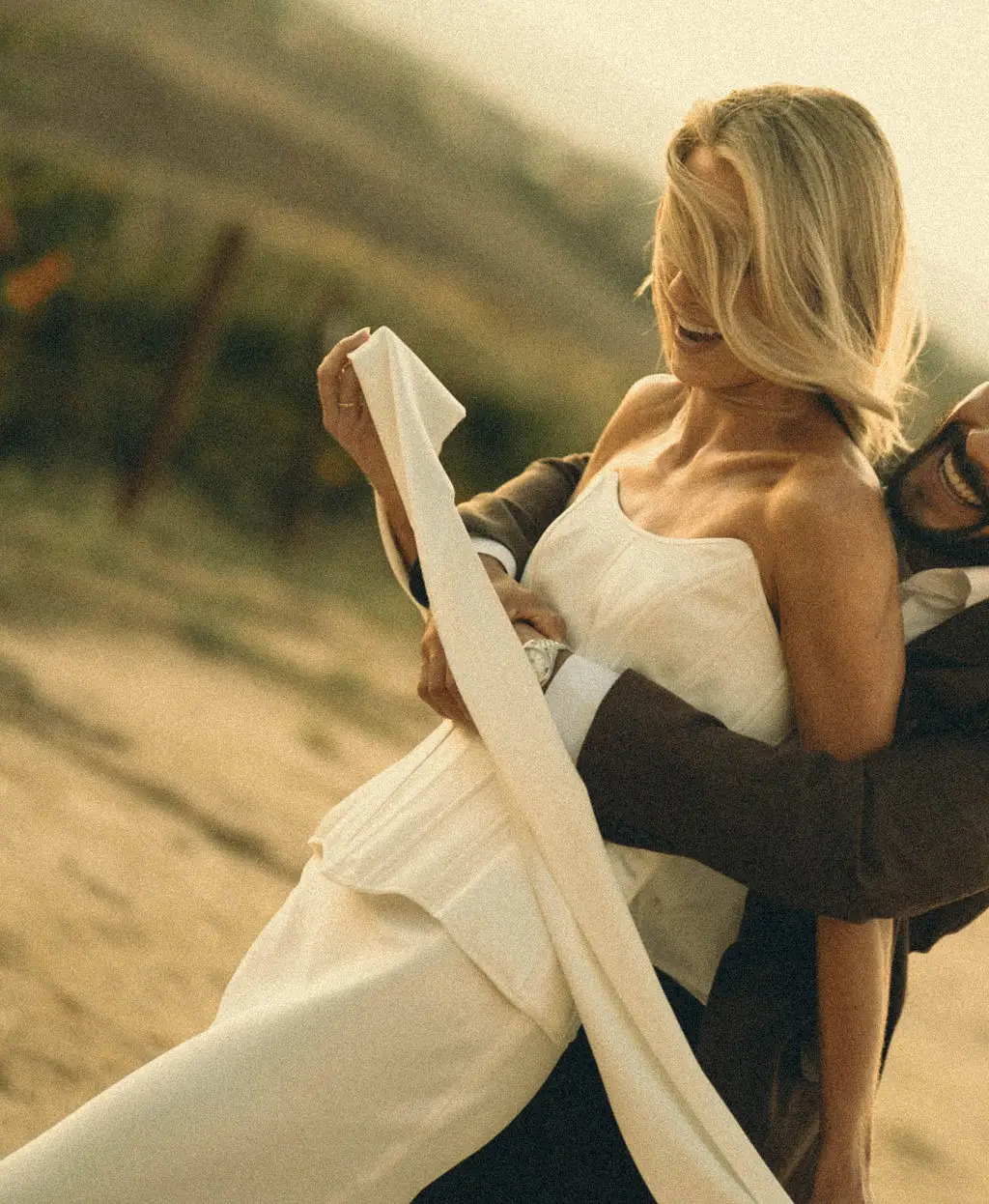 Groom lifts laughing bride against golden hillside light at a warm outdoor wedding