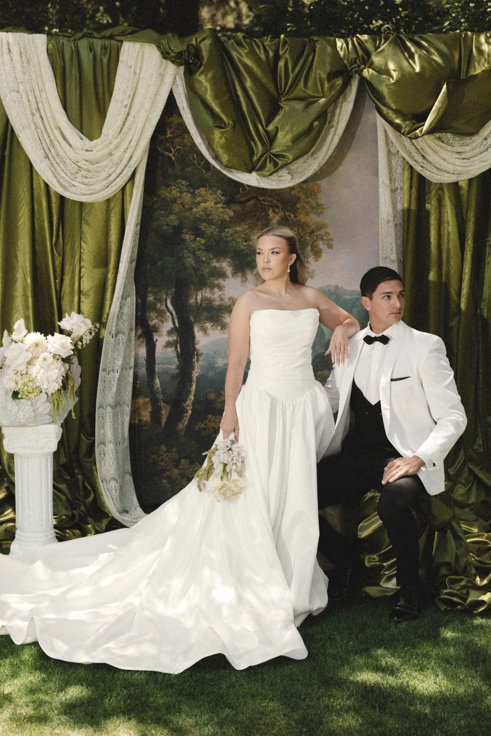 Bride with a long flowing train and groom in a tuxedo beneath an olive-draped wedding arch at Havens Country Club in Vista, California.