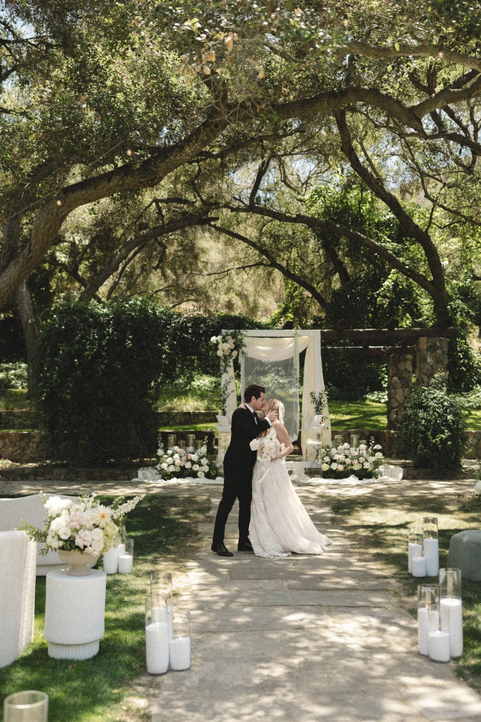Wedding ceremony arch draped in sheer fabric at the end of an oak-lined garden path at Havens Country Club in Vista, California.