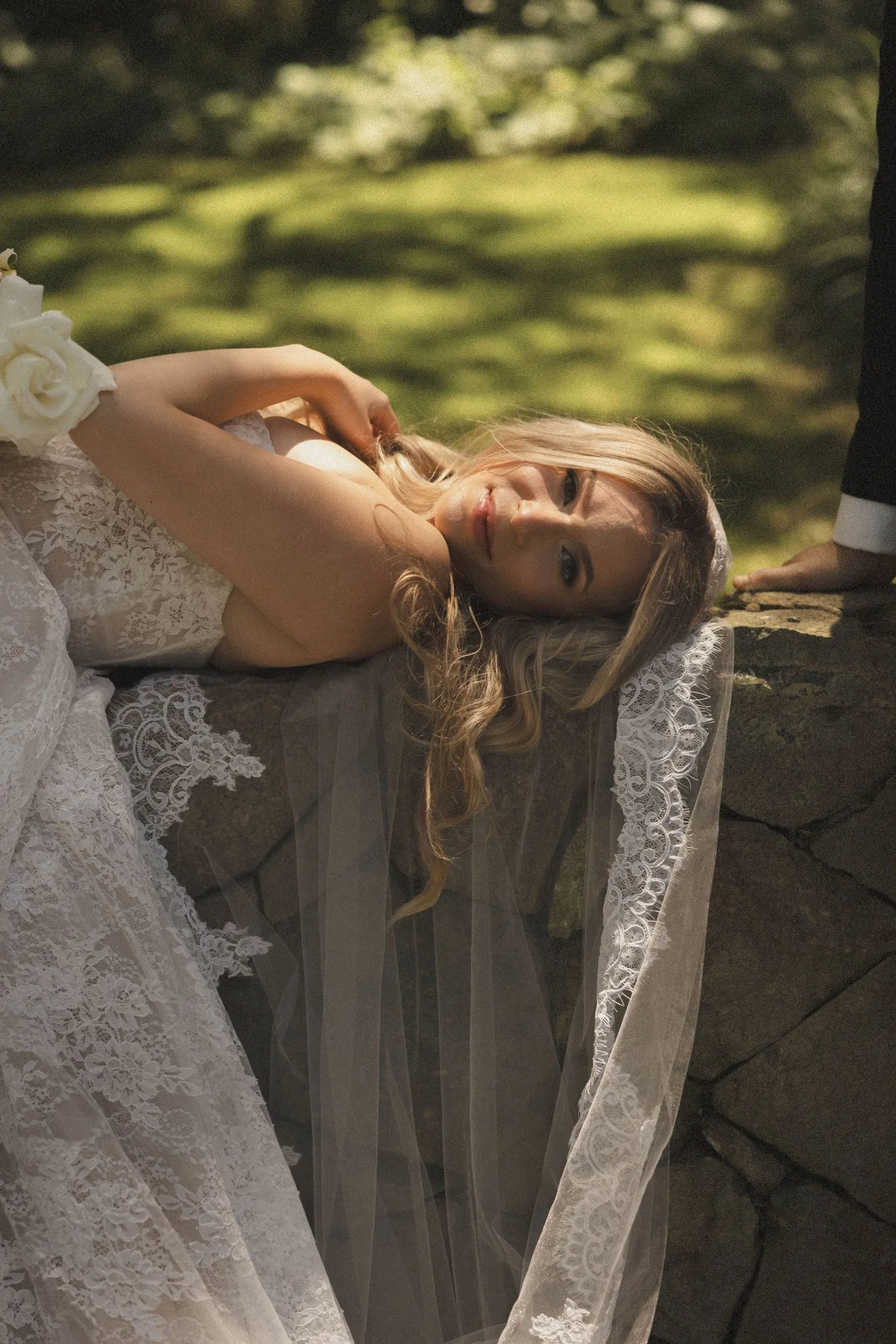 Bride in a lace-trimmed veil resting against a stone wall in dappled sunlight at Havens Country Club, an editorial wedding portrait.