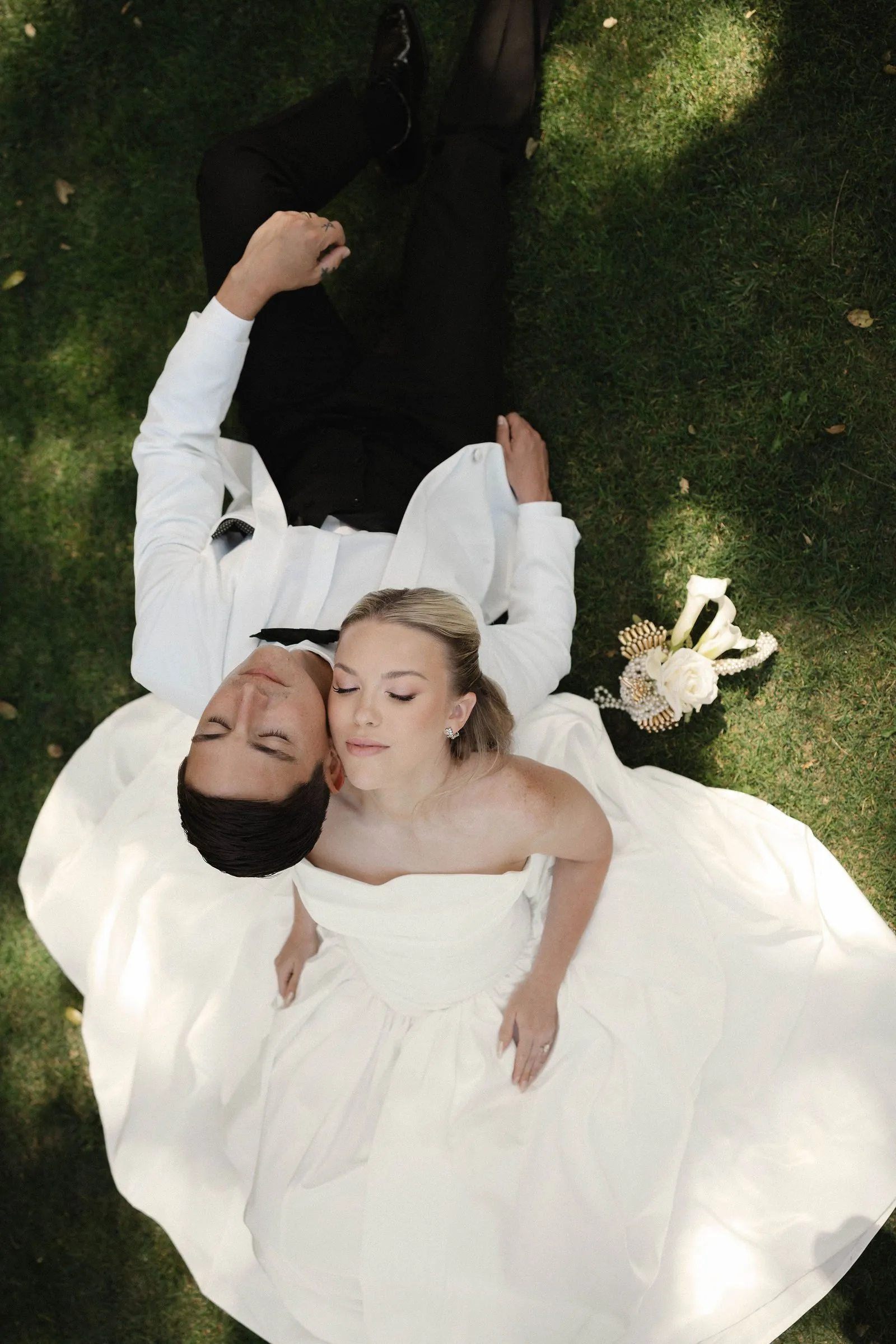 Bride and groom lying on the lawn at Havens Country Club photographed from above, her gown and bouquet spread across the grass.