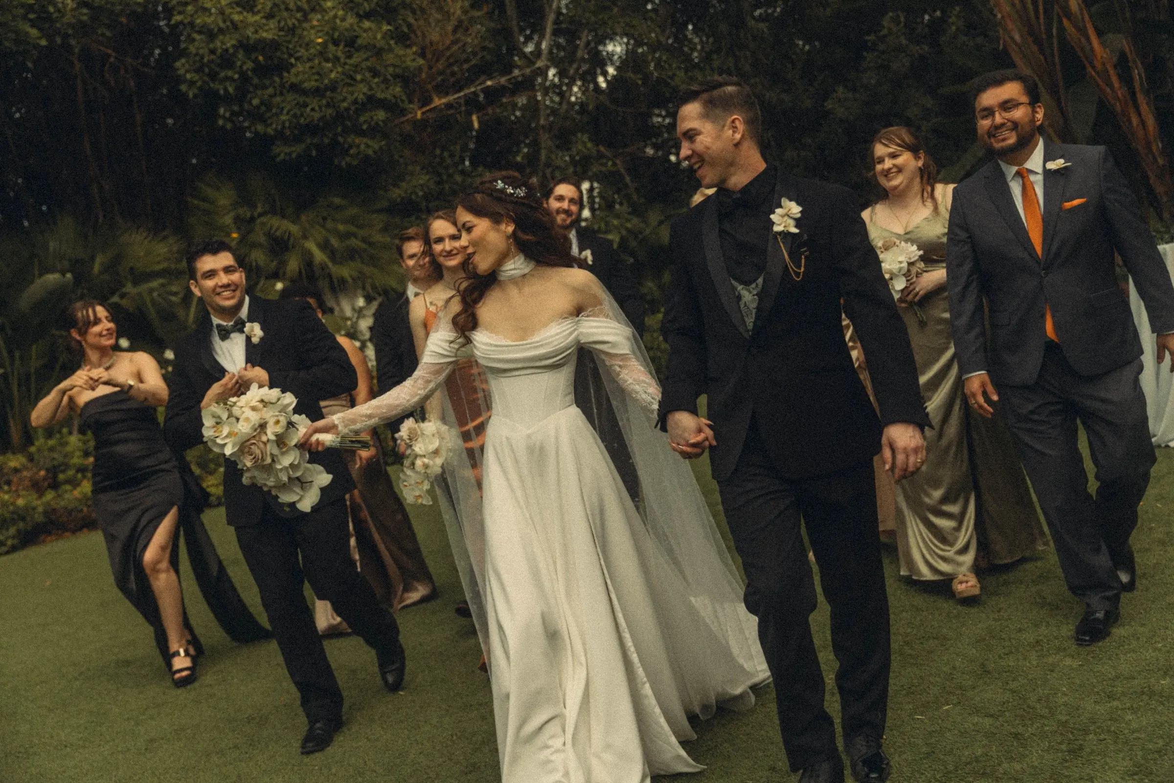 Bride and groom walk hand in hand with their wedding party across a garden lawn.