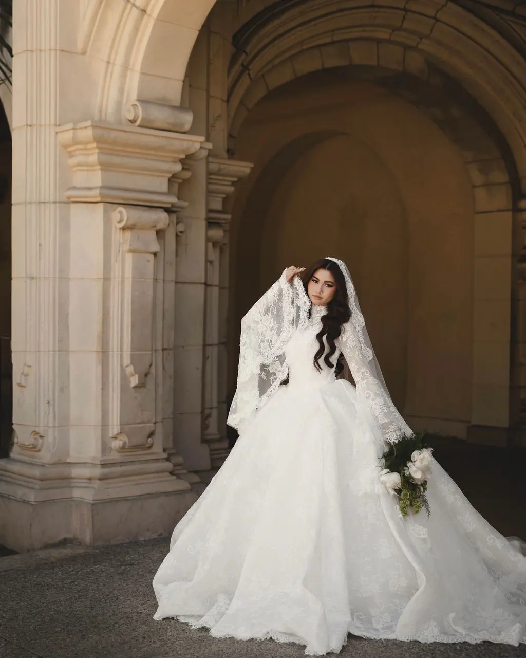 Bride in a lace ballgown and veil lifts her veil beneath a grand stone archway and columns.